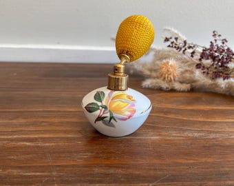 Könnte beinhalten: Eine Vintage-Parfümflasche mit weißem Sockel, verziert mit einem gelb-rosa Rosenmuster. Sie hat einen goldfarbenen Zerstäuber und einen strukturierten gelben Ball. Die Flasche ist ein elegantes Dekorationsstück.