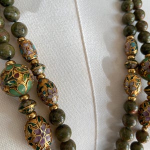 Puede incluir: Un collar de doble cadena con cuentas de piedra verde y cuentas de esmalte verde, dorado y blanco con dise&ntilde;os florales.