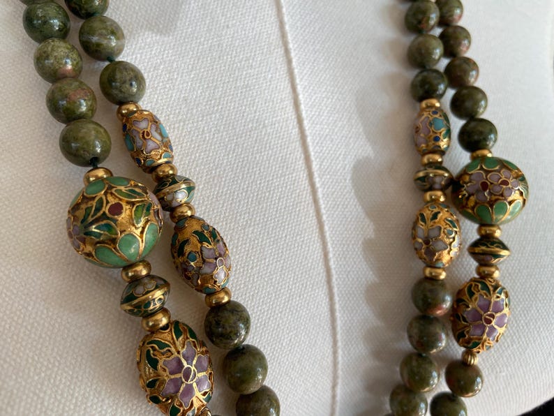 Puede incluir: Un collar de doble cadena con cuentas de piedra verde y cuentas de esmalte verde, dorado y blanco con dise&ntilde;os florales.