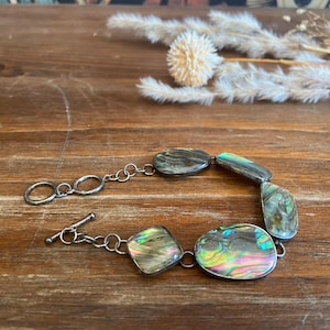 May include: Abalone shell bracelet with iridescent colors and a silver chain. The bracelet features oval and irregular-shaped abalone shell pieces linked together with a toggle clasp. The bracelet is displayed on a wooden surface.
