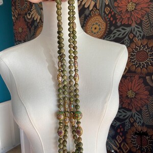 Puede incluir: Un collar de m&uacute;ltiples hilos hecho de cuentas verdes y marrones con detalles dorados. Las cuentas son redondas y tienen un patr&oacute;n moteado. El collar es largo y llega hasta el pecho.