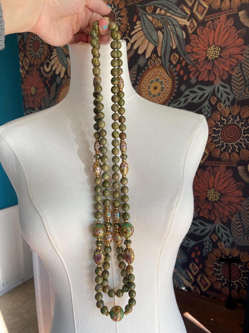 Puede incluir: Un collar de m&uacute;ltiples hilos hecho de cuentas verdes y marrones con detalles dorados. Las cuentas son redondas y tienen un patr&oacute;n moteado. El collar es largo y llega hasta el pecho.