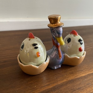 May include: A vintage ceramic salt and pepper shaker set featuring a duck wearing a top hat and scarf, flanked by two chicks emerging from eggshells. The duck is purple with a gold hat, and the chicks are white with red accents.