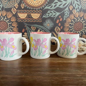 May include: Three white ceramic mugs with a pink interior and a floral iris design in pink, yellow, and green. The mugs have a curved handle and are set against a patterned background.
