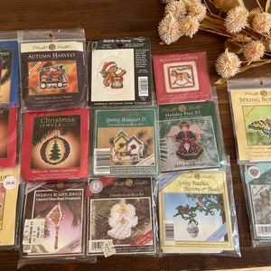 May include: A collection of 14 counted cross stitch and bead kits featuring various holiday and seasonal themes. The kits include designs such as a rocking horse, a Christmas tree, a garden cart, a joyful angel, and a spring bouquet. The kits are packaged in clear plastic bags with colourful labels and images of the finished designs.