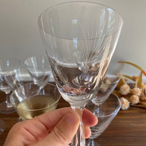 May include: Clear stemmed wine glasses with etched floral designs. The glasses have a slender stem and a flared bowl. Several glasses are visible, some filled with a light-colored liquid. The background includes dried flowers and a wooden surface.
