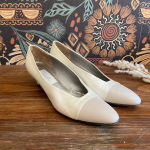May include: A pair of off-white women's pumps with a light gray toe cap. The shoes have a low heel and a glossy finish. The shoes are displayed on a wooden surface.
