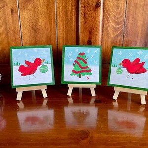 以下が含まれることがあります： ミニイーゼルに飾られた3つの小さなクリスマスをテーマにした絵画。2つの絵画は、緑のオーナメントを運ぶ赤い鳥を描いています。3つ目は、装飾されたクリスマスツリーを描いています。すべての絵画は、緑色の縁と水色の背景を持っています。