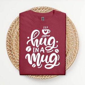 Op de afbeelding: Een maroon t-shirt met witte tekst die "hug in a mug" luidt met een koffiekopje afbeelding boven de tekst.