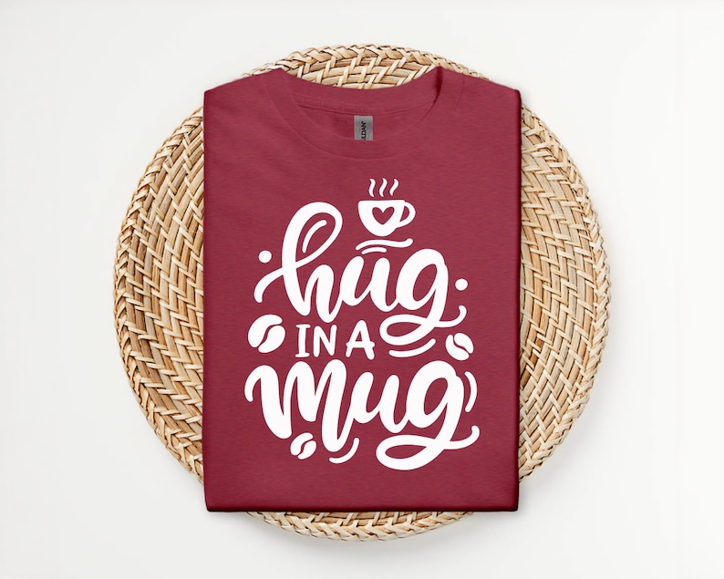 Op de afbeelding: Een maroon t-shirt met witte tekst die "hug in a mug" luidt met een koffiekopje afbeelding boven de tekst.