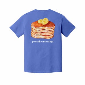 Pu&ograve; includere: Maglietta blu con un'illustrazione di una pila di pancake con fette di banana. La frase "pancake mornings" &egrave; stampata in bianco sotto. La maglietta &egrave; di colore blu unito.