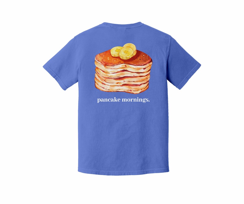 Pu&ograve; includere: Maglietta blu con un'illustrazione di una pila di pancake con fette di banana. La frase "pancake mornings" &egrave; stampata in bianco sotto. La maglietta &egrave; di colore blu unito.