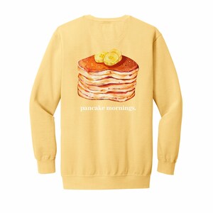 May include: Yellow long-sleeve sweatshirt with a graphic of a stack of pancakes topped with banana slices. The text "pancake mornings." is printed below the image.
