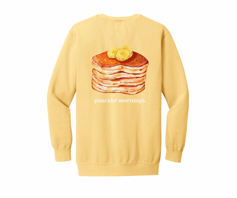 May include: Yellow long-sleeve sweatshirt with a graphic of a stack of pancakes topped with banana slices. The text "pancake mornings." is printed below the image.