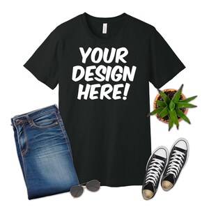 K&ouml;nnte beinhalten: Ein schwarzes T-Shirt mit dem Text "YOUR DESIGN HERE!" in Wei&szlig;. Das T-Shirt liegt flach auf einem wei&szlig;en Hintergrund mit einer blauen Jeans, einer Sonnenbrille und einer Topfpflanze.