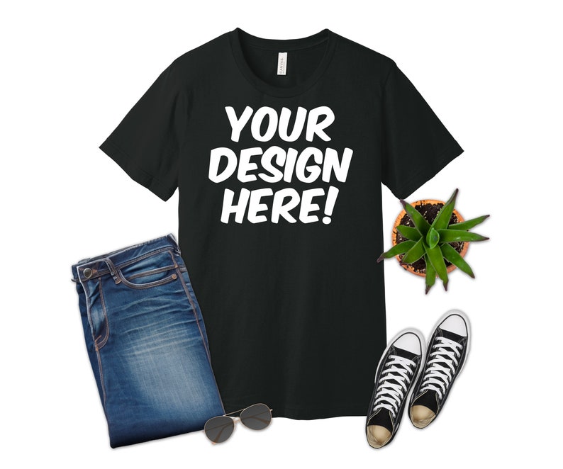 K&ouml;nnte beinhalten: Ein schwarzes T-Shirt mit dem Text "YOUR DESIGN HERE!" in Wei&szlig;. Das T-Shirt liegt flach auf einem wei&szlig;en Hintergrund mit einer blauen Jeans, einer Sonnenbrille und einer Topfpflanze.
