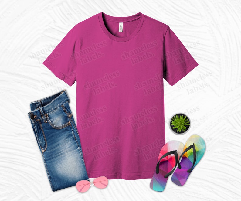 K&ouml;nnte beinhalten: Ein pinkfarbenes T-Shirt mit kurzen &Auml;rmeln und dem Text "shameless labels." wiederholt auf der Vorderseite. Das Shirt liegt flach auf einer wei&szlig;en Oberfl&auml;che mit einer blauen Jeans, einem Paar Regenbogen-Flip-Flops und einer kleinen Topfpflanze.