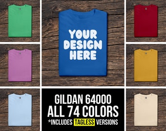 Op de afbeelding: Een mockup van een Gildan 64000 T-shirt in verschillende kleuren. Het T-shirt is gevouwen en wordt weergegeven op een houten oppervlak. De tekst "YOUR DESIGN HERE" is gedrukt op het blauwe T-shirt. De tekst "GILDAN 64000 ALL 74 COLORS *INCLUDES TAGLESS VERSIONS" wordt weergegeven onder de T-shirts.