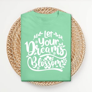 Op de afbeelding: Een lichtgroen T-shirt met de tekst "Let your dreams blossom" in witte letters met bloemmotieven.