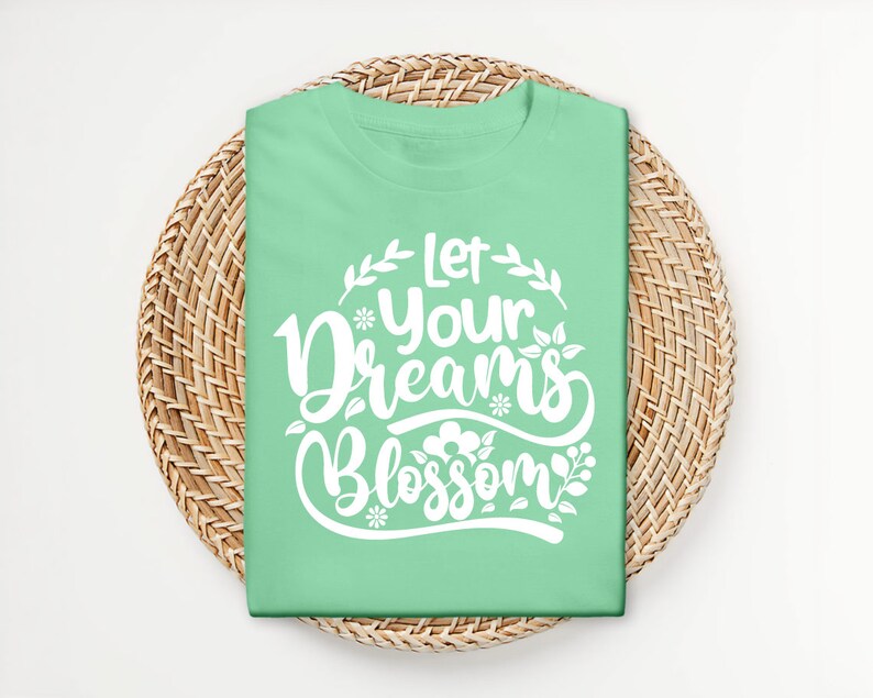 Op de afbeelding: Een lichtgroen T-shirt met de tekst "Let your dreams blossom" in witte letters met bloemmotieven.