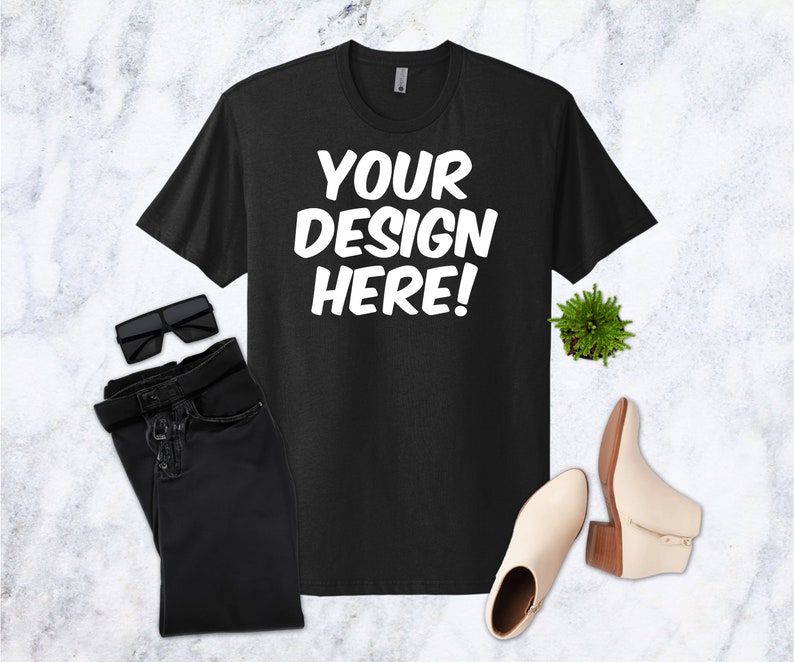 May include: Black t-shirt with the text "YOUR DESIGN HERE!" in white, paired with black sunglasses, black jeans, a small green plant, and cream-colored ankle boots with a wooden heel.