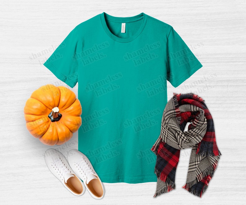 K&ouml;nnte beinhalten: Ein t&uuml;rkisfarbenes T-Shirt mit einem wei&szlig;en K&uuml;rbis, zwei wei&szlig;en Turnschuhen und einem schwarz-rot karierten Schal auf einem wei&szlig;en Holzgrund.