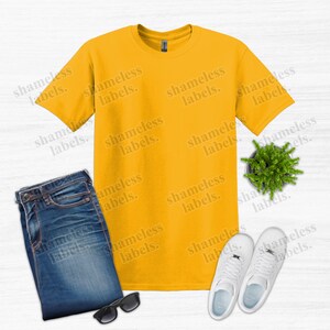 K&ouml;nnte beinhalten: Draufsicht mit einem senfgelben T-Shirt, gefalteten blauen Jeans, einer schwarzen Sonnenbrille, einer kleinen Topfpflanze und wei&szlig;en Turnschuhen. Der Text "shameless labels" ist als Wasserzeichen im Bild.