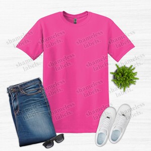 K&ouml;nnte beinhalten: Ein leuchtend pinkfarbenes T-Shirt, blaue Jeans, wei&szlig;e Turnschuhe, eine Sonnenbrille und eine kleine Topfpflanze sind auf einer hellen Holzmaserung angeordnet. Der Text "shameless labels" ist schwach sichtbar.