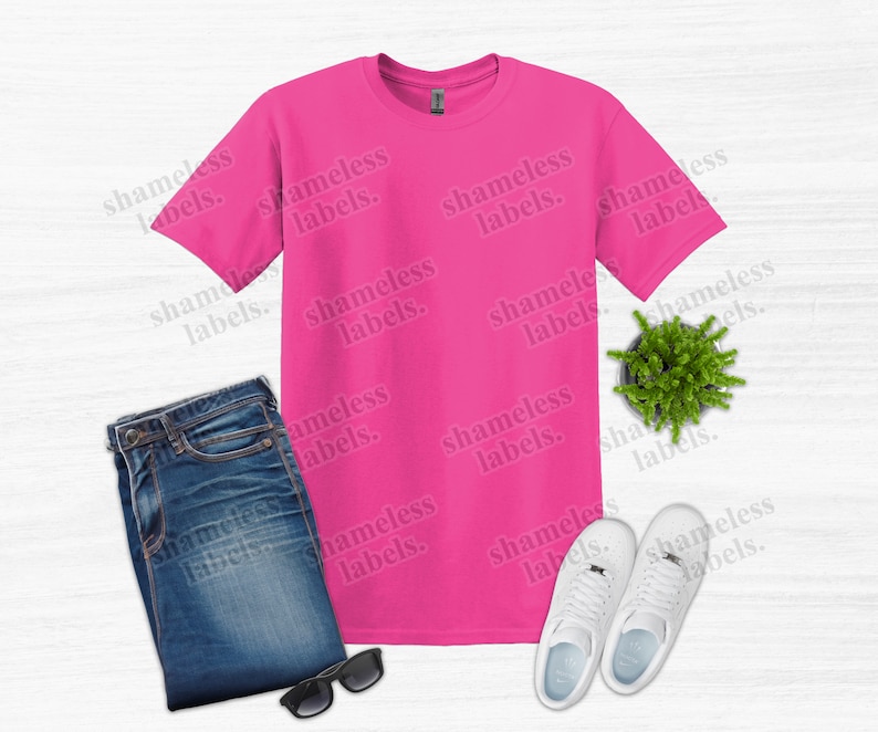 K&ouml;nnte beinhalten: Ein leuchtend pinkfarbenes T-Shirt, blaue Jeans, wei&szlig;e Turnschuhe, eine Sonnenbrille und eine kleine Topfpflanze sind auf einer hellen Holzmaserung angeordnet. Der Text "shameless labels" ist schwach sichtbar.