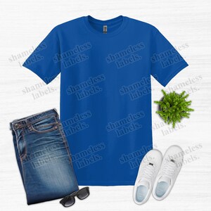 K&ouml;nnte beinhalten: Ein k&ouml;nigsblaues T-Shirt, gefaltete Jeans, eine schwarze Sonnenbrille, wei&szlig;e Turnschuhe und eine kleine Topfpflanze sind auf einer wei&szlig;en Oberfl&auml;che angeordnet. Auf dem T-Shirt steht der Text "shameless labels".