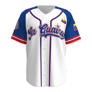 Puede incluir: Jersey de béisbol blanco con ribetes azules y rojos. El jersey tiene una estrella azul y el texto "La Guaira" en una fuente azul cursiva. La bandera venezolana está en la manga derecha.