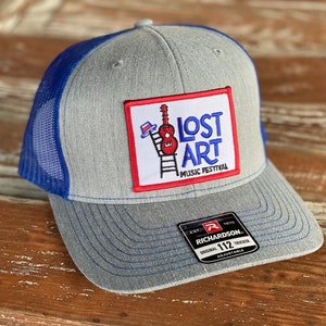 Puede incluir: Gorra de camionero gris y azul con un parche rojo, blanco y azul que dice "Lost Art Music Festival". El parche presenta una ilustración de dibujos animados de una guitarra, un sombrero y una escalera. La gorra es una Richardson 112.