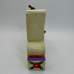 May include: A carved wooden figurine with a cream-colored body, a purple base, and a red flower design. The figurine has a small red and black detail on top. The base features a red flower with green leaves.