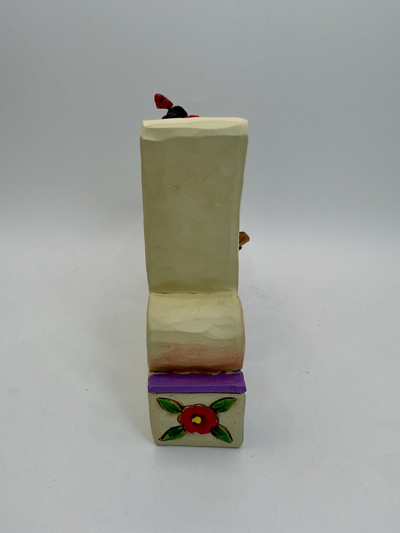 May include: A carved wooden figurine with a cream-colored body, a purple base, and a red flower design. The figurine has a small red and black detail on top. The base features a red flower with green leaves.