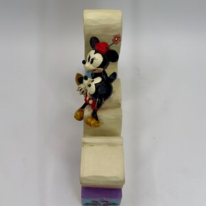 May include: A decorative figurine featuring Minnie Mouse and Mickey Mouse climbing a staircase. Minnie Mouse wears a red hat with a flower, a red and white polka dot skirt, and yellow shoes. Mickey Mouse wears a blue shirt and white gloves. The staircase is beige with a purple and blue base.