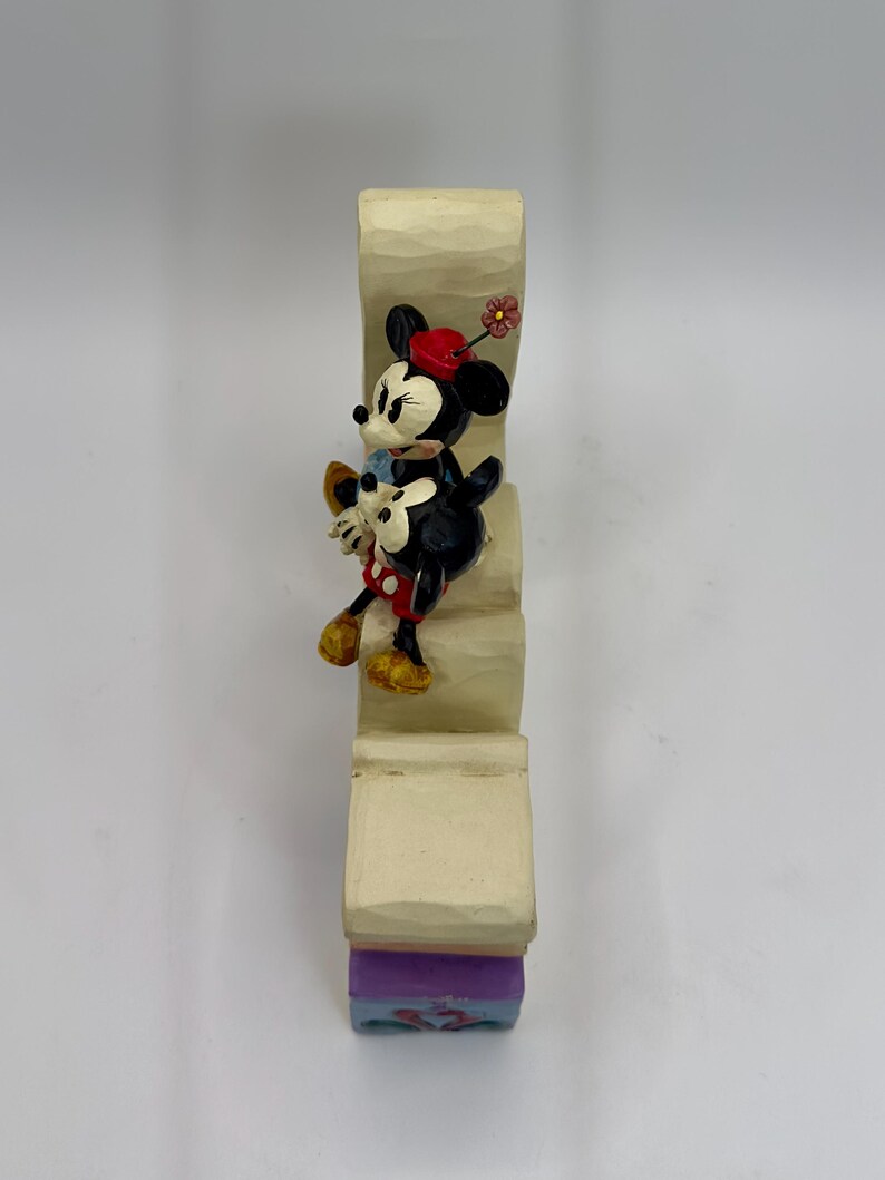 May include: A decorative figurine featuring Minnie Mouse and Mickey Mouse climbing a staircase. Minnie Mouse wears a red hat with a flower, a red and white polka dot skirt, and yellow shoes. Mickey Mouse wears a blue shirt and white gloves. The staircase is beige with a purple and blue base.