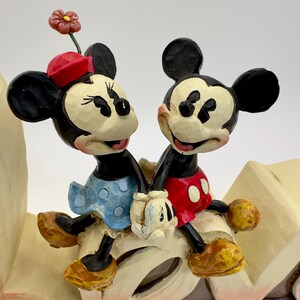 May include: A figurine of Mickey and Minnie Mouse holding hands. Minnie wears a red hat and blue polka dot dress. Mickey wears red shorts with white buttons. The figurine is on a decorative base with floral accents.