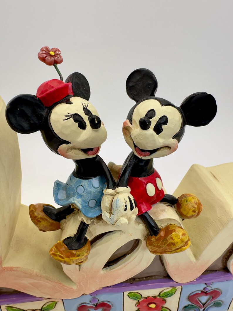 May include: A figurine of Mickey and Minnie Mouse holding hands. Minnie wears a red hat and blue polka dot dress. Mickey wears red shorts with white buttons. The figurine is on a decorative base with floral accents.