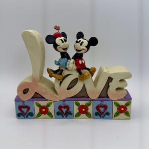 May include: A decorative figurine featuring Mickey and Minnie Mouse sitting atop the word "LOVE". The base is decorated with hearts and flowers. The figurine is painted in soft pastel colors.