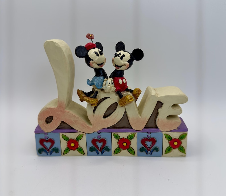 May include: A decorative figurine featuring Mickey and Minnie Mouse sitting atop the word "LOVE". The base is decorated with hearts and flowers. The figurine is painted in soft pastel colors.