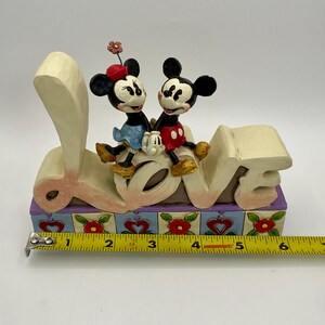 May include: A decorative figurine featuring Mickey and Minnie Mouse sitting on top of the word "LOVE". The figurine has a colorful base with floral and heart designs. Mickey is wearing red shorts, and Minnie is wearing a blue dress.