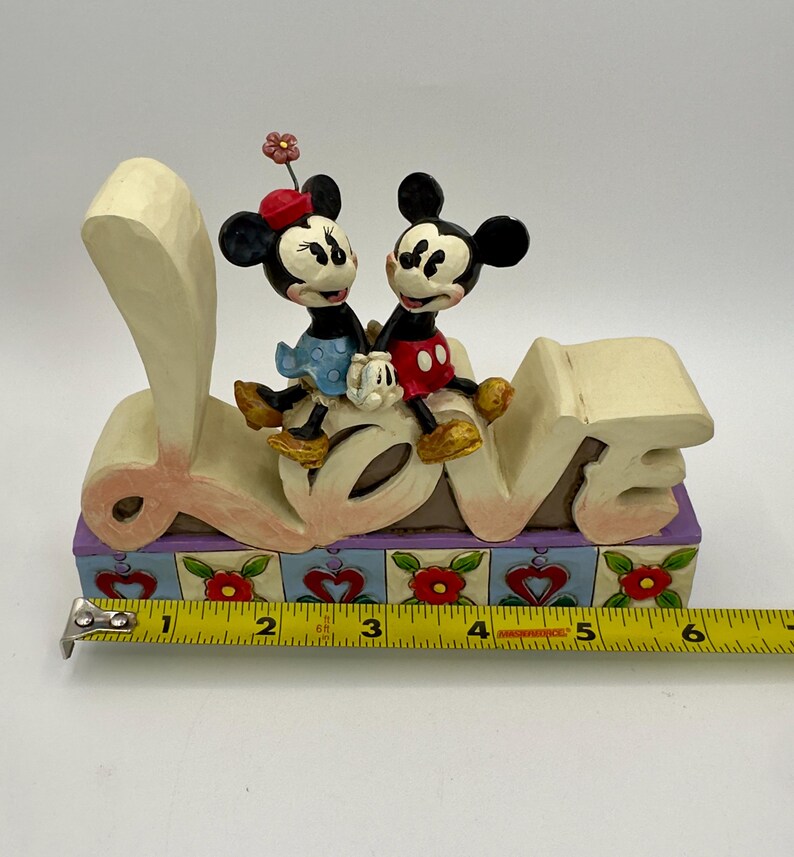May include: A decorative figurine featuring Mickey and Minnie Mouse sitting on top of the word "LOVE". The figurine has a colorful base with floral and heart designs. Mickey is wearing red shorts, and Minnie is wearing a blue dress.