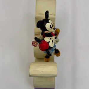 May include: A decorative figurine featuring Mickey Mouse and Minnie Mouse climbing a light-colored structure. Mickey is in a red shirt and black shorts, while Minnie wears a red dress and holds a flower. The structure has a purple base.