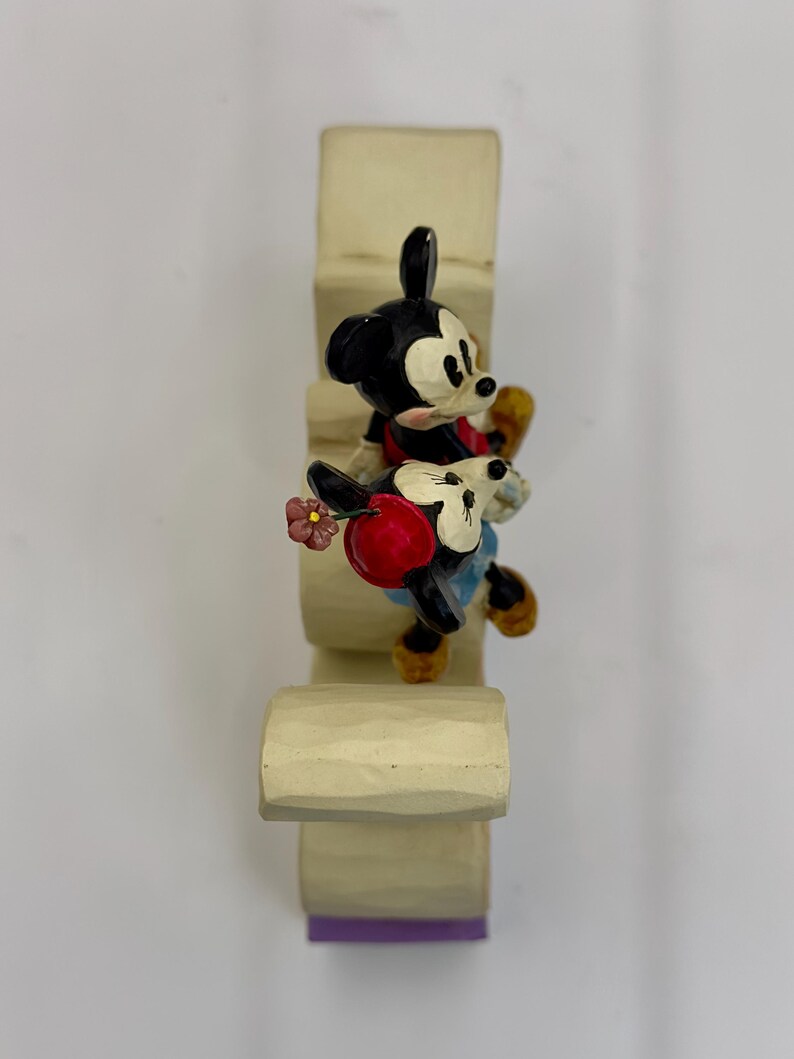 May include: A decorative figurine featuring Mickey Mouse and Minnie Mouse climbing a light-colored structure. Mickey is in a red shirt and black shorts, while Minnie wears a red dress and holds a flower. The structure has a purple base.