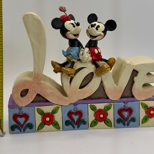 May include: A decorative figurine featuring Mickey and Minnie Mouse sitting atop the word "LOVE". The base is adorned with hearts and flowers. The figurine measures approximately 4 inches in height.