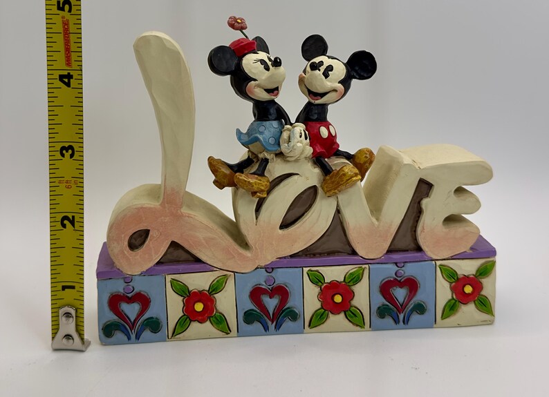 May include: A decorative figurine featuring Mickey and Minnie Mouse sitting atop the word "LOVE". The base is adorned with hearts and flowers. The figurine measures approximately 4 inches in height.