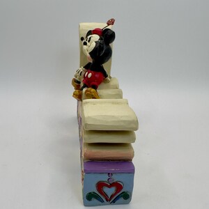 May include: A decorative figurine featuring Mickey Mouse in a seated pose atop a multi-tiered, colorful block. Mickey is wearing his signature red shorts, yellow shoes, and a red hat. The block is painted with pastel colors and heart designs.