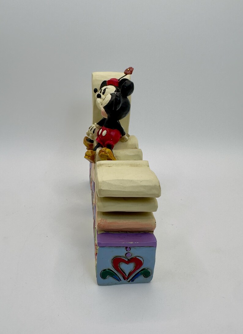 May include: A decorative figurine featuring Mickey Mouse in a seated pose atop a multi-tiered, colorful block. Mickey is wearing his signature red shorts, yellow shoes, and a red hat. The block is painted with pastel colors and heart designs.