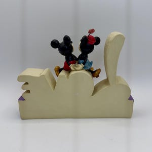 May include: A cream-colored decorative figurine featuring Mickey and Minnie Mouse figures seated on a stylized "LOVE" base. Mickey wears red shorts, and Minnie wears a red hat and blue skirt. The base has purple accents.