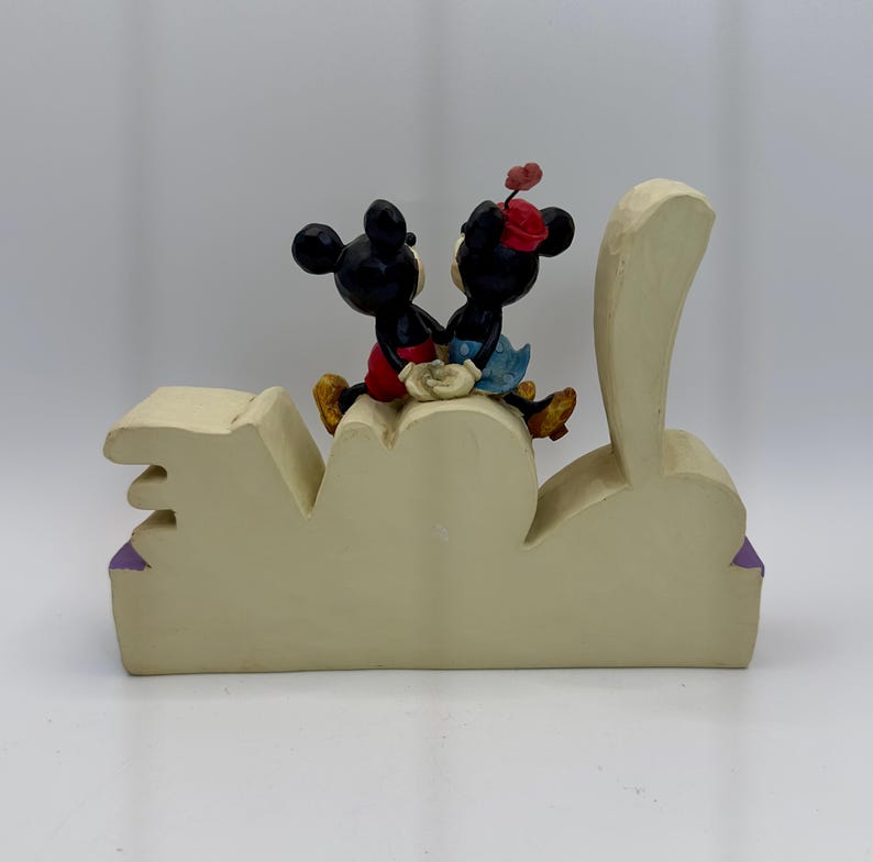 May include: A cream-colored decorative figurine featuring Mickey and Minnie Mouse figures seated on a stylized "LOVE" base. Mickey wears red shorts, and Minnie wears a red hat and blue skirt. The base has purple accents.